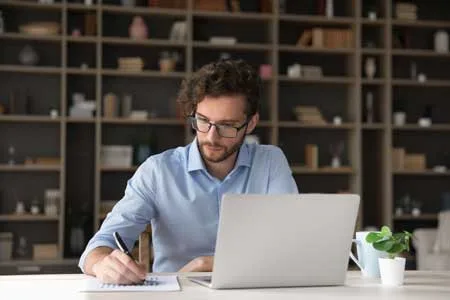 Student sitting at desk in front of laptop writing in notebook